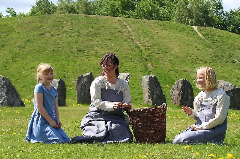 Barn och pedagog sitter framför Anundshögen. Annonsbild för Järnåldersliv 2023. Foto Nina Larsen